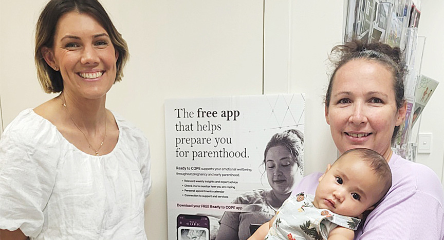 Nurse Olivia Renwick and new mother Casey Watkins, holding baby Amanki. They are smiling and standing in front of a poster about 'Ready to COPE' - a free app that helps prepare you for parenthood.