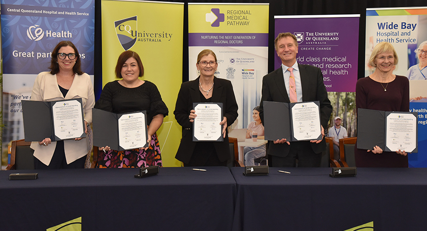 Associate Professor Catherine McDougall (Chief Medical Officer, Queensland Health), Lisa Blackler (Chief Executive, Central Queensland Hospital and Health Service), Debbie Carroll (Chief Executive, Wide Bay Hospital and Health Service), Professor Nick Klomp (Vice-Chancellor and President, CQUniversity Australia) and Professor Deborah Terry AC (Vice-Chancellor and President, The University of Queensland) standing behind a table each holding up signed Memorandum of Understanding documents for the Regional Medical Pathway. In the background are promotional banners for each organisation.