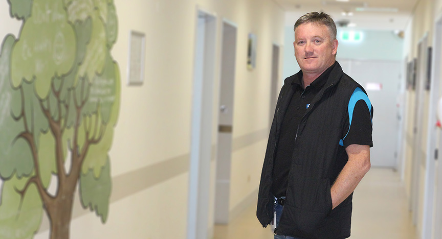 Clinical Nurse Consultant Mark stands in a hallway with a tree mural on the wall