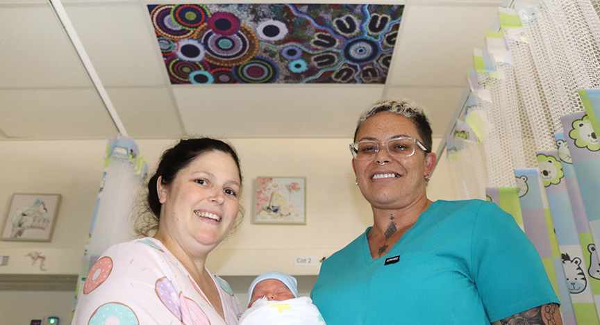 Smiling mum Misty holds baby Rip, wrapped in a bunny rug and wearing a beanie. Connie stands beside them wearing glasses and a teal scrub top. Above them is ceiling tile featuring a multicoloured indigenous artwork.