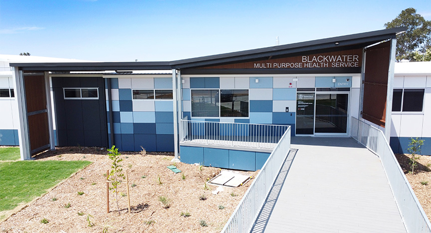 exterior photo of a newly constructed multipurpose health service, featuring timber accents, panels of different shades of blue, a large entry ramp, new turf and newly planted gardens