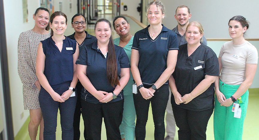 A group of nine allied health clinicians and support staff stand for a team photo in a health clinic hallway with green lino flooring and lots of natural light.