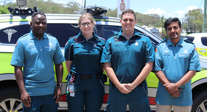 Douglas, Kayla, Max and Partha wear their QAS uniforms while standing in front of an ambulance vehicle.