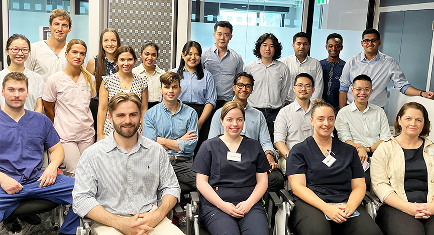 Group of 21 young medical graduates sitting and standing in a group, wearing a mix of hospital scrubs and business attire, in the training room of the hospital academic centre