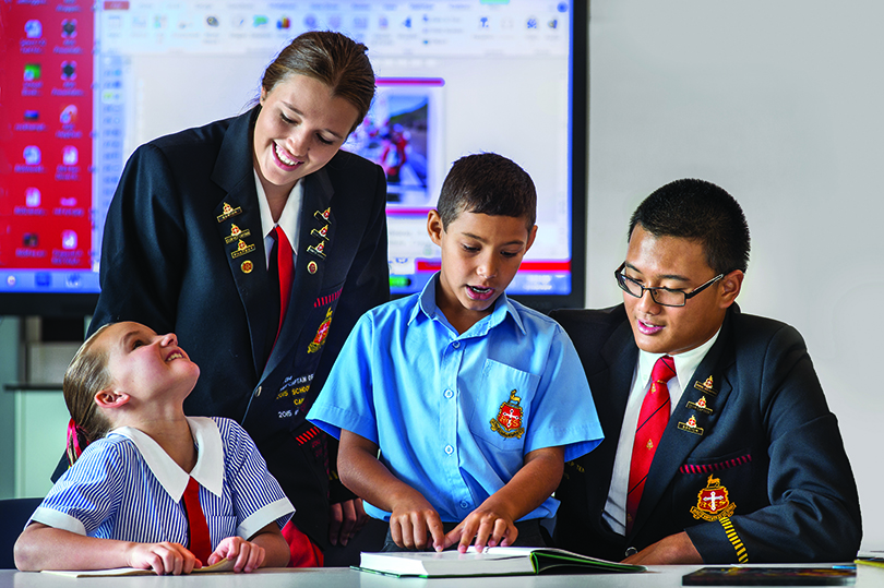 Four school students gather around a desk. Two of the students are reading books.