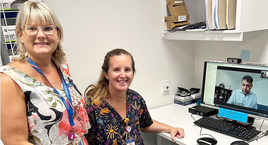 Chontal is wearing glasses and a floral dress and is standing next to Emily, who is sitting at a computer wearing black scrubs with a multi-coloured floral pattern. Dr Gupta is on the computer screen wearing a pale blue business shirt.