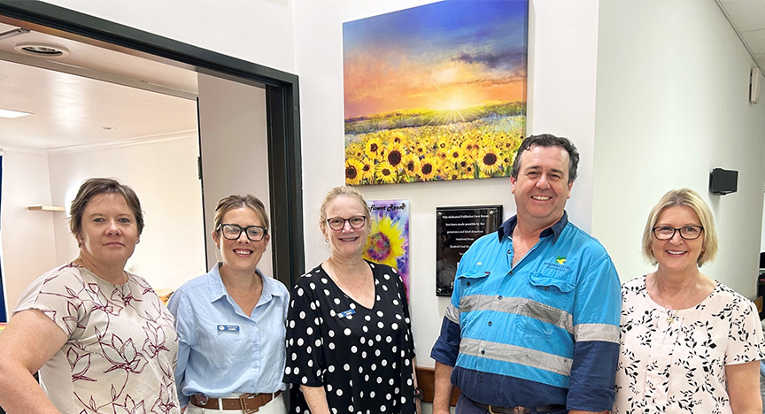 Four women wearing office attire and a man in hi-vis workwear stand outside the new sunflower palliative care room, in front of a canvas of sunflowers at sunset and a plaque recognising donations from Kestrel Coal.