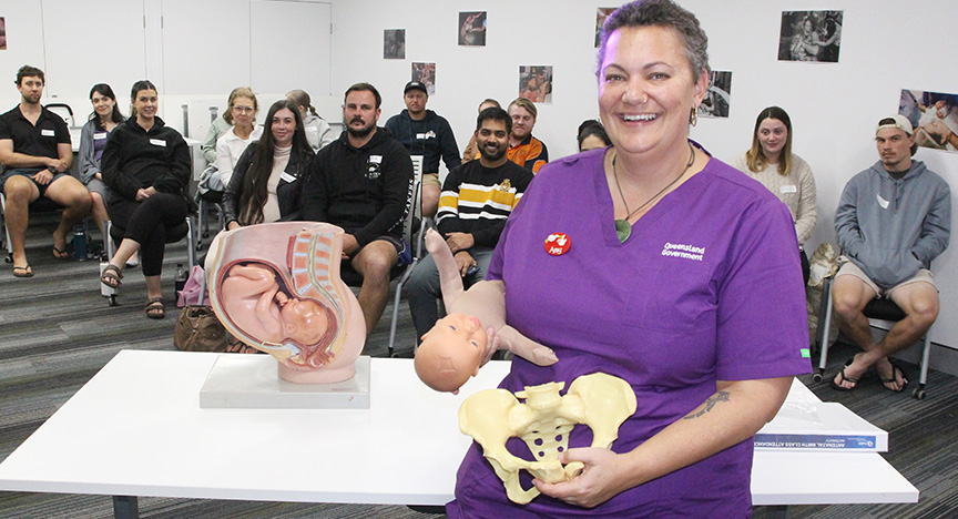 Midwife wearing purple scrubs smiles for the camera. She's holding a skeleton model of a pelvis and a baby mannequin, standing in front of a table with an education model of a womb and baby. In the background, a crowd of men and women are seated to watch the presentation.