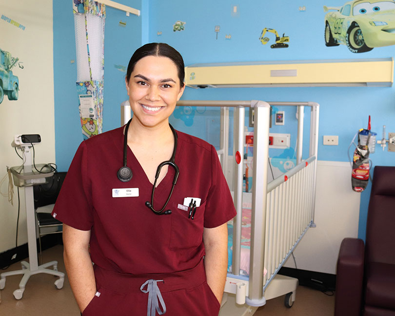 Doctor wearing a stethoscope around neck smiles at the camera. In the background is a paediatric ward bed and cartoon stickers on the wall.