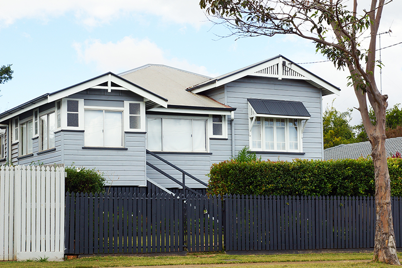 A two-storey Queenslander house surrounded by well-maintained hedges and a dark grey fence.