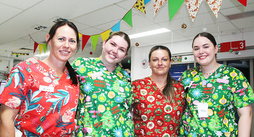 Four nurses stand together in the Emergency Department wearing bright red and green Christmas scrubs and surrounded by Christmas decorations.