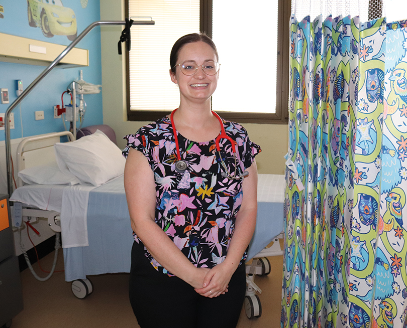 A smiling doctor from CQ Health sits in front of a hospital bed. A stethoscope hangs around their neck.