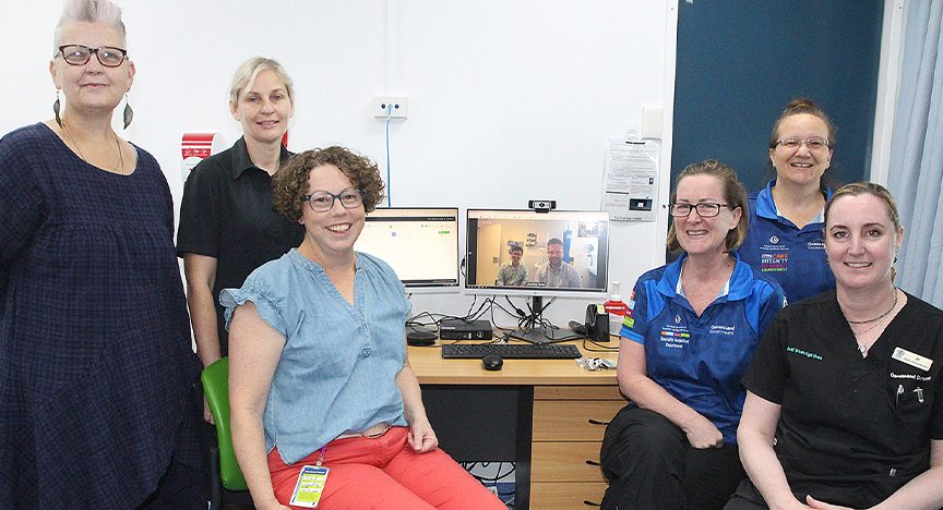 Group of healthcare workers gather around a computer in a hospital clinic with two specialist doctors on the screen dialling in via telehealth