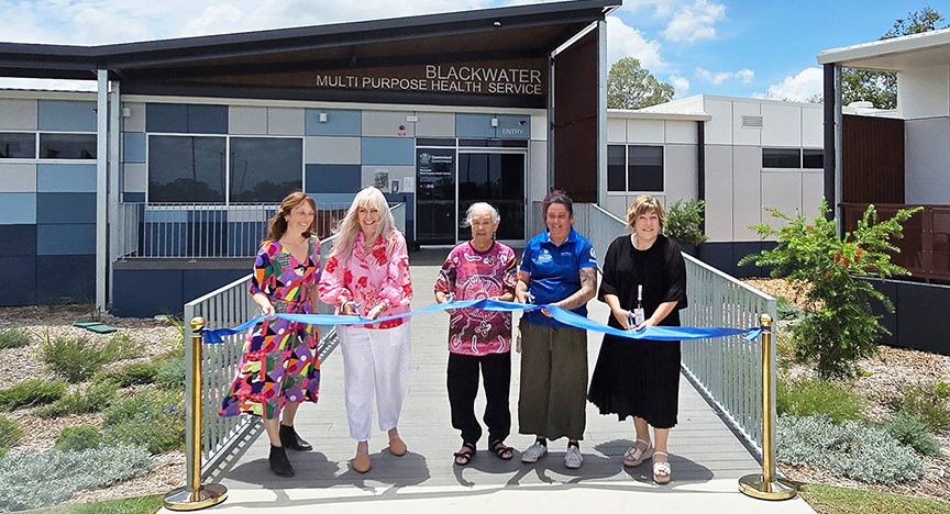 Four women stand ready to cut a long blue ribbon between two gold-coloured bollards outside the front entrance of the hospital with the words Blackwater Multipurpose Health Service on a dark brown gable.