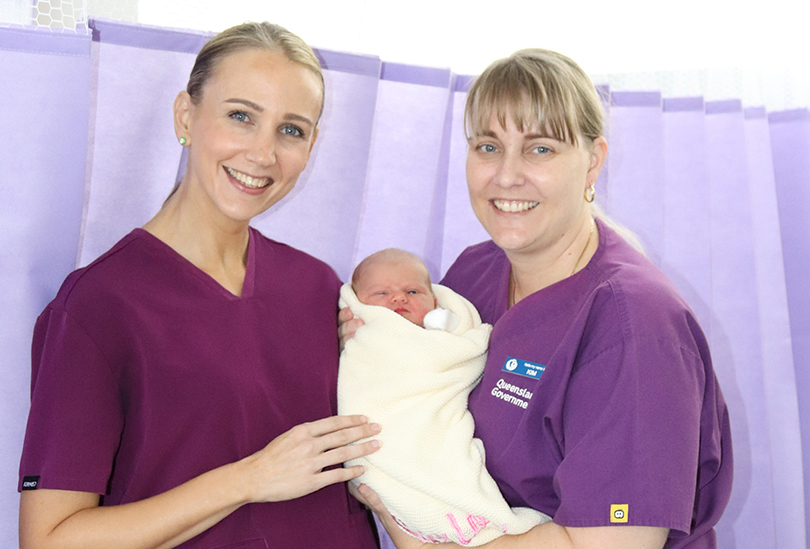 Two midwives wearing purple scrubs smile at the camera. One of them is holding a baby wrapped in a blanket. They are standing in front of a purple curtain divider in a maternity ward.